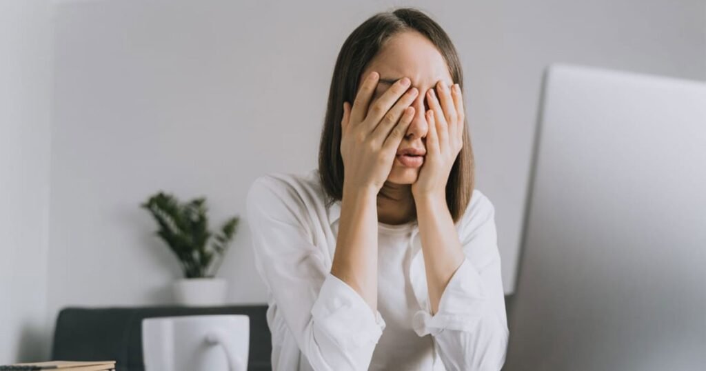 A woman at a desk, shielding her eyes with her hands, indicating a moment of eye strain or fatigue.